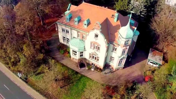 Un grande edificio decorato con numerose torri e un tetto rosso, immerso nel verde e con un viale lastricato.