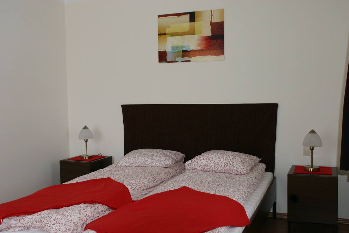 Una cama bien tendida con una manta roja, dos mesitas de noche con lámparas y una obra de arte en la pared sobre la cama.