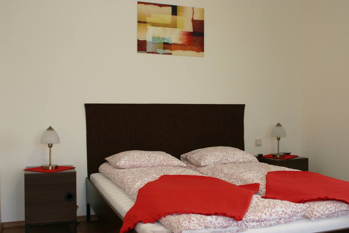 Una cama bien hecha con una colcha roja, dos mesitas de noche con lámparas y una obra de arte en la pared sobre la cama.