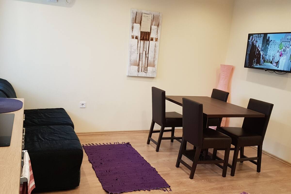 Una habitación pequeña y moderna con una mesa de comedor de madera, cuatro sillas, una alfombra morada, un reposapiés negro y un televisor montado en la pared.