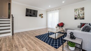 A modern living room with a staircase, a flat-screen TV, a sofa, a coffee table, and a painting of the Eiffel Tower. - Entire Bungalow 5 Bedrooms (Markham)