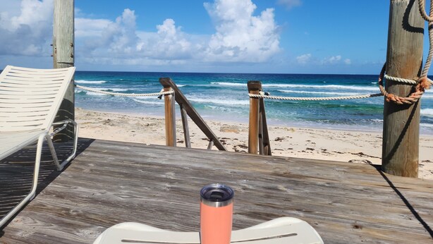Une terrasse en bois avec un fauteuil de jardin blanc et une rambarde en métal, donnant sur une plage avec des vagues et un ciel dégagé.