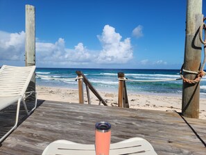 A wooden deck with a white lounge chair and a metal railing, overlooking a beach with waves and a clear sky. - Oceanfront 2BR, Villa | Private Beach, HotTub Starlink +Generator
 (Long Island)
