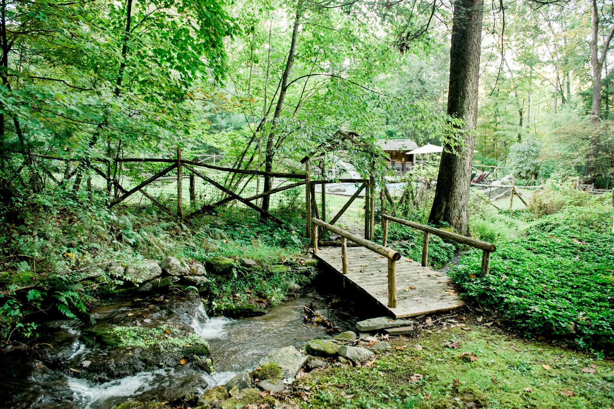 A wooden bridge over a stream in a forested area with a building in the background.