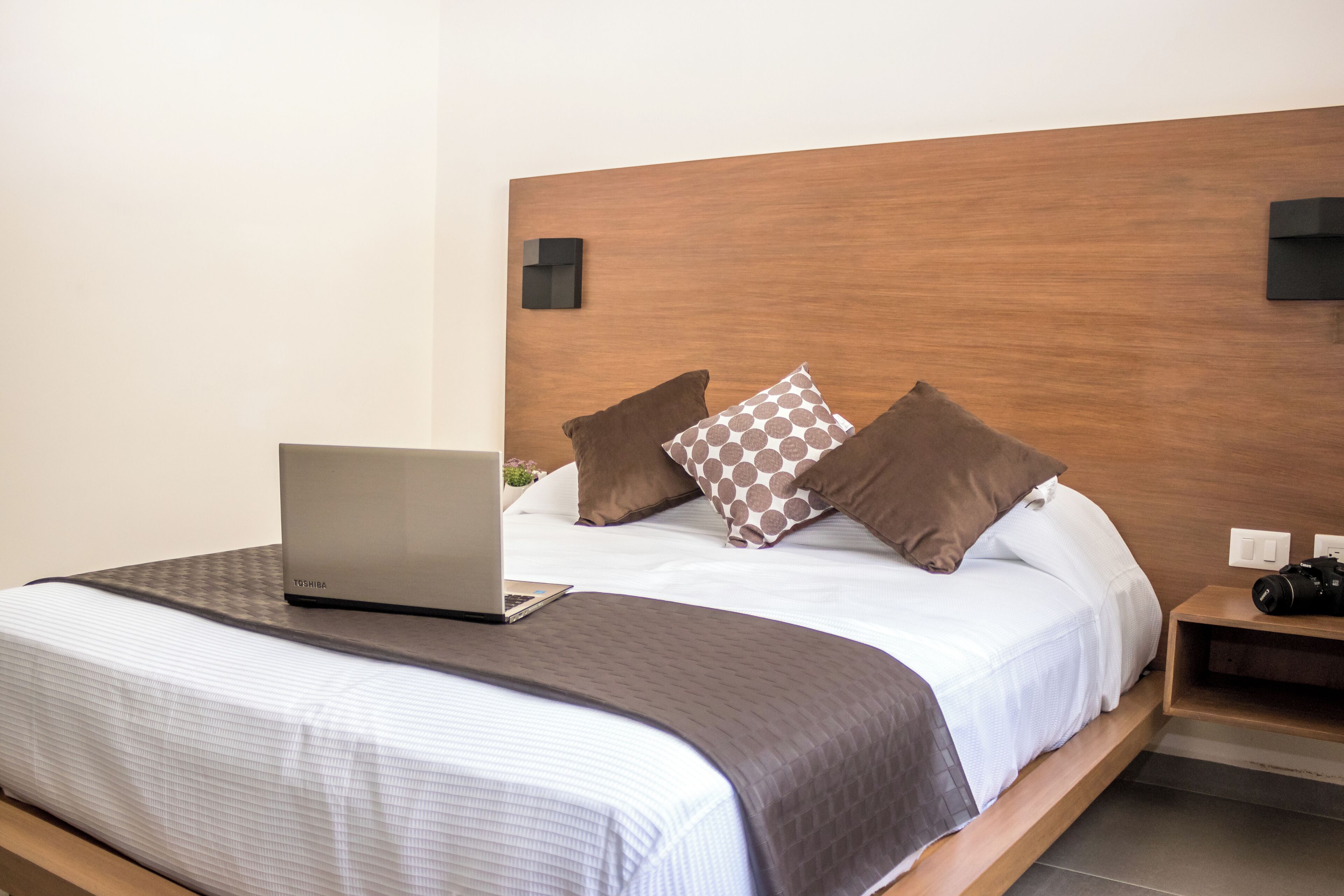 Een net opgemaakt bed met een laptop, kussens en een nachtkastje met een camera.