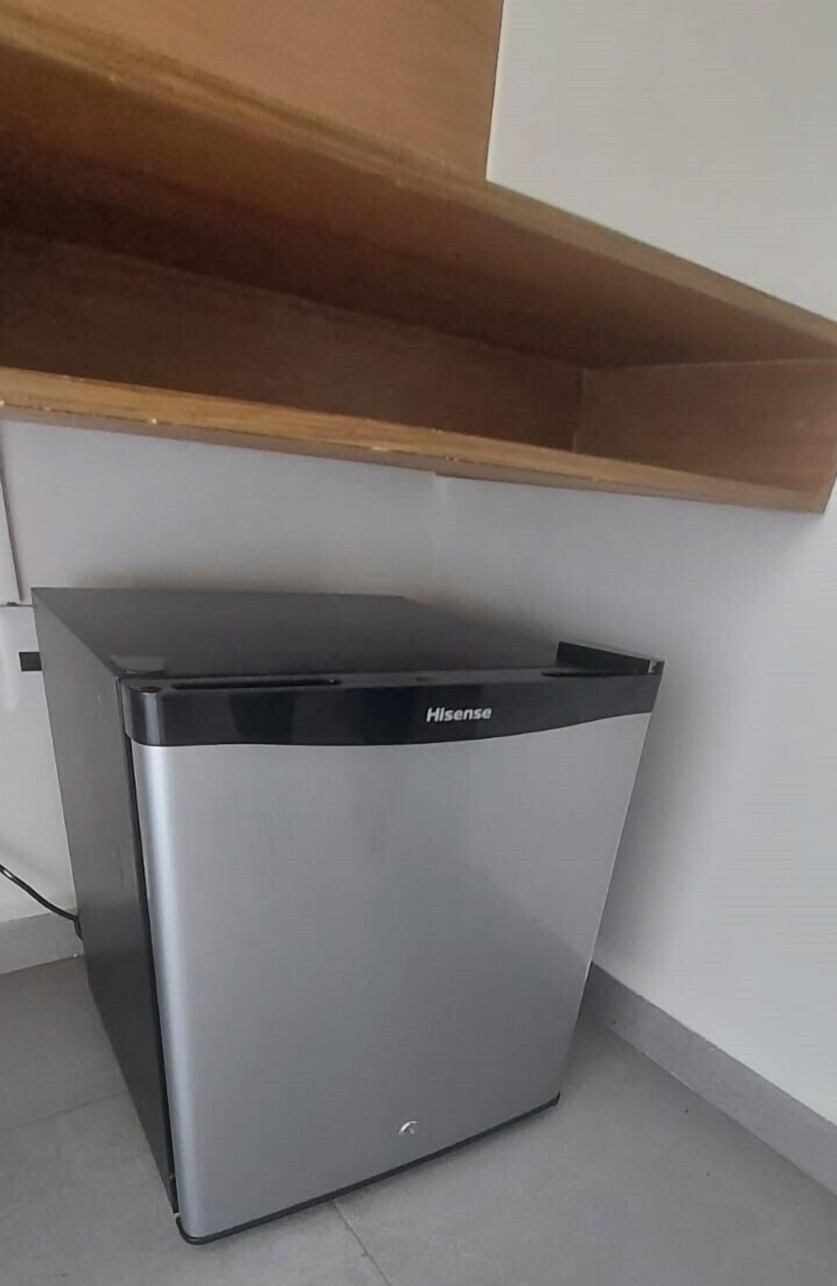 A compact, stainless steel trash bin with a black lid, labeled "Hisense," placed in a corner under a wooden shelf.