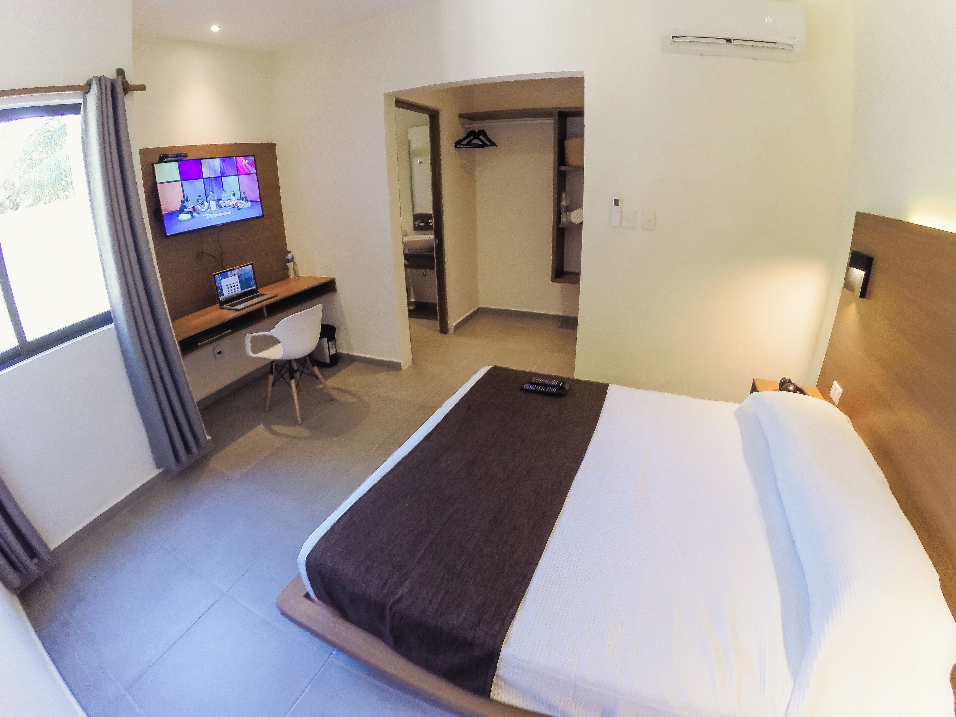 A hotel room with a bed, a desk with a laptop, a TV, and a bathroom visible through an open door.