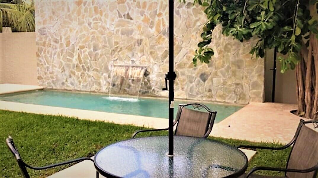 An outdoor pool area with a stone wall, a small waterfall feature, and a round glass table with chairs.