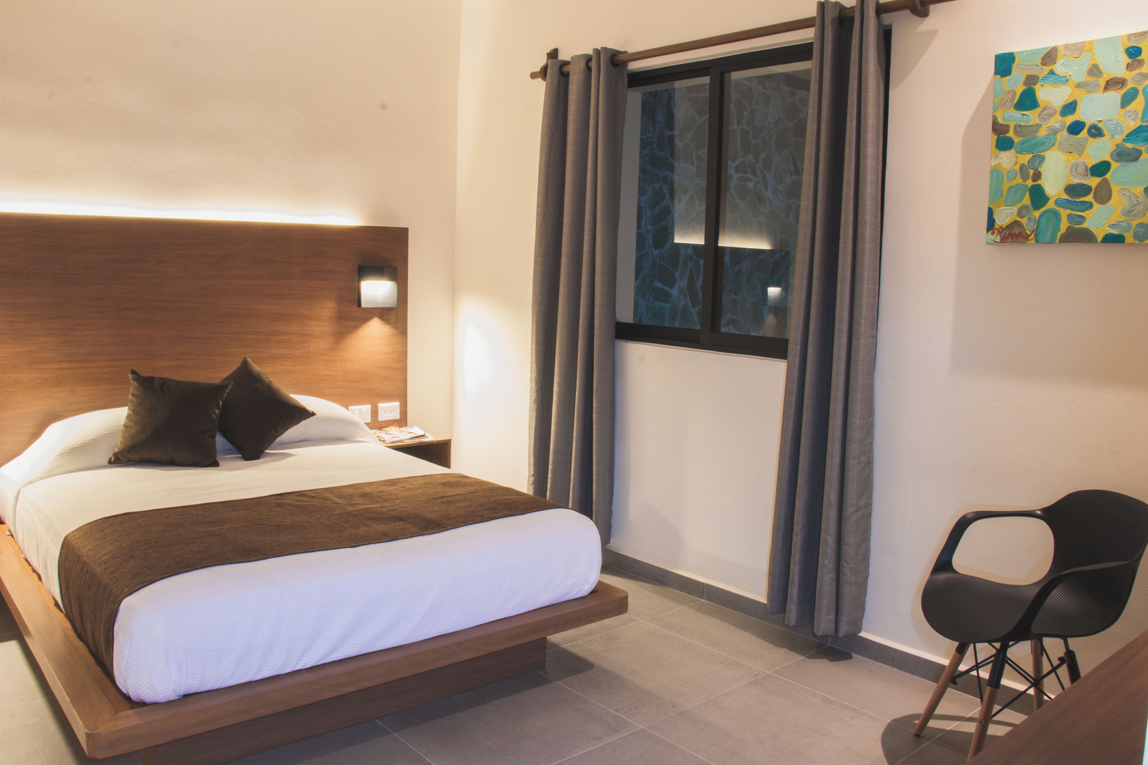 Quarto de hotel moderno com uma cama grande, mesa de cabeceira, cadeira e janela com cortinas.