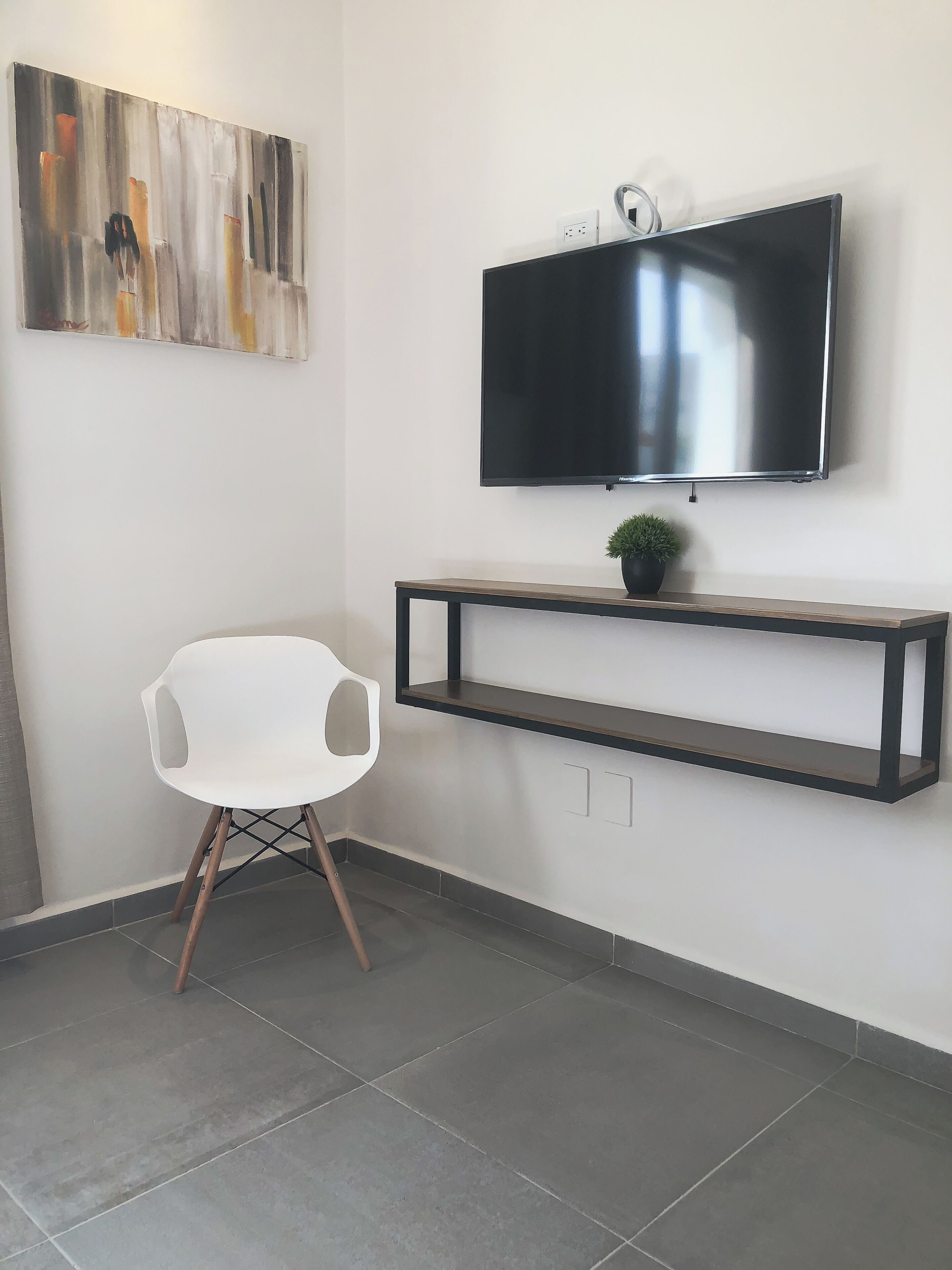 Una habitación moderna con un televisor de pantalla plana montado en la pared, una silla blanca, un estante minimalista y una pequeña planta en maceta.
