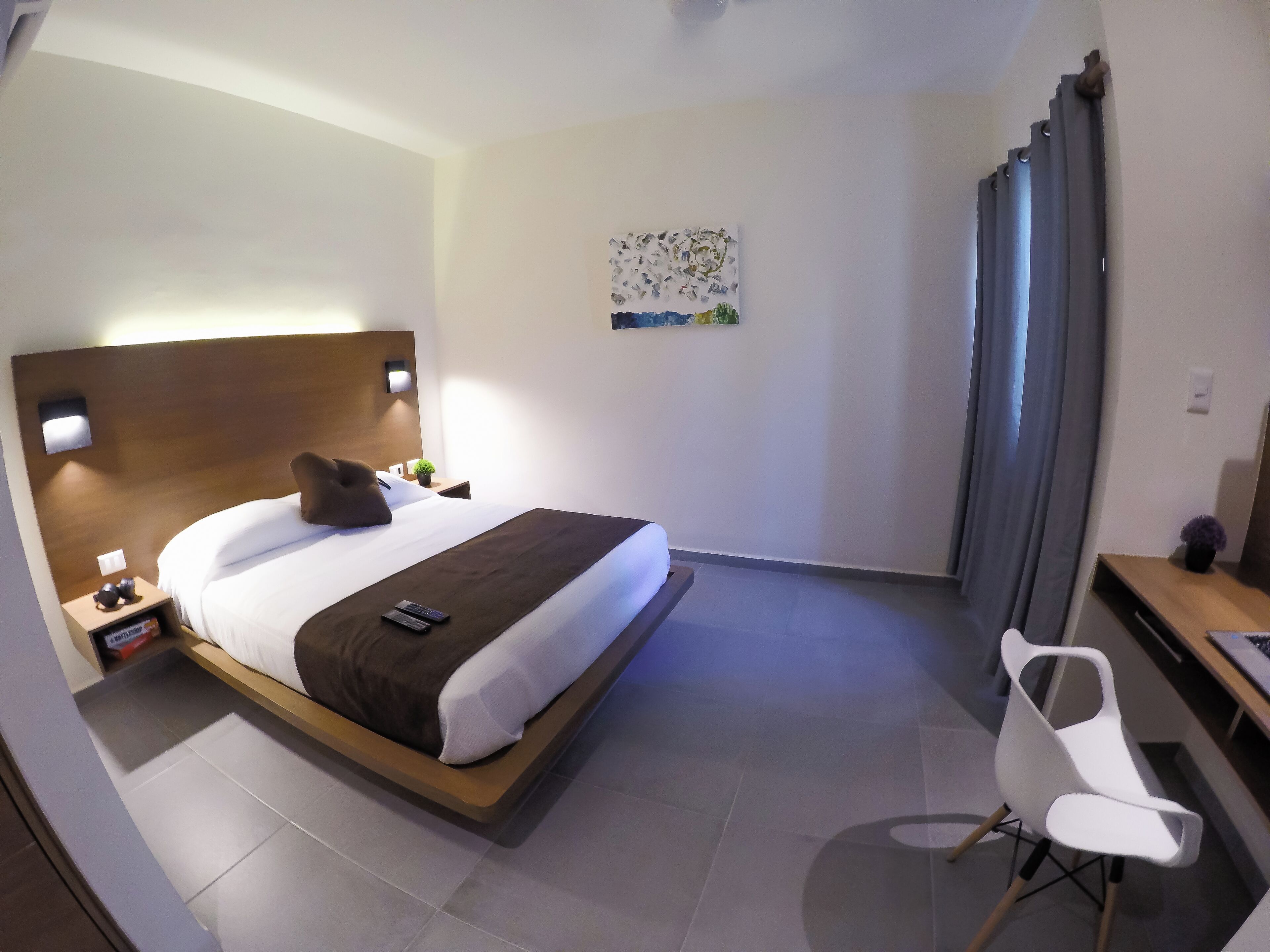 A modern hotel room with a large bed, bedside table, lamp, and a desk with a chair.