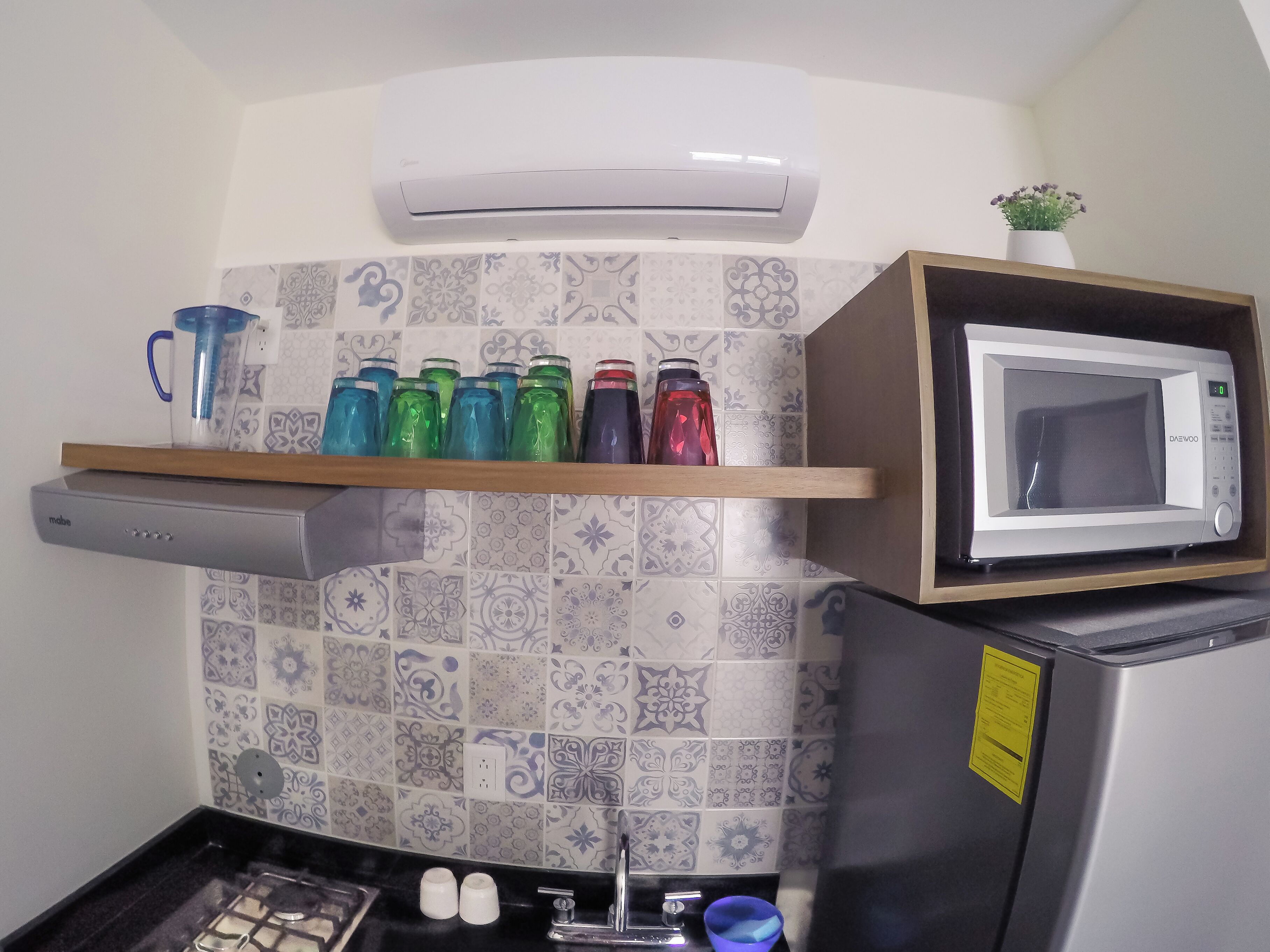 Une cuisine équipée d’un four à micro-ondes, d’une étagère avec des bouteilles d’eau de différentes couleurs, d’une cuisinière à gaz et d’un climatiseur fixé au mur.