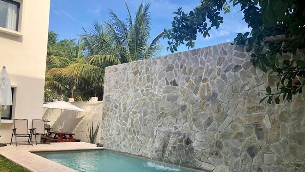 Una piscina con un muro in pietra, un patio con sedie e una palma sullo sfondo.