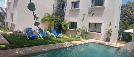 A modern building with a swimming pool, blue lounge chairs, and potted plants.