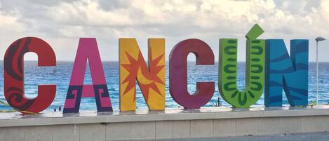 A colorful sign spelling 'CANCUN' with a stylized design, located near a waterfront with a view of the sea.