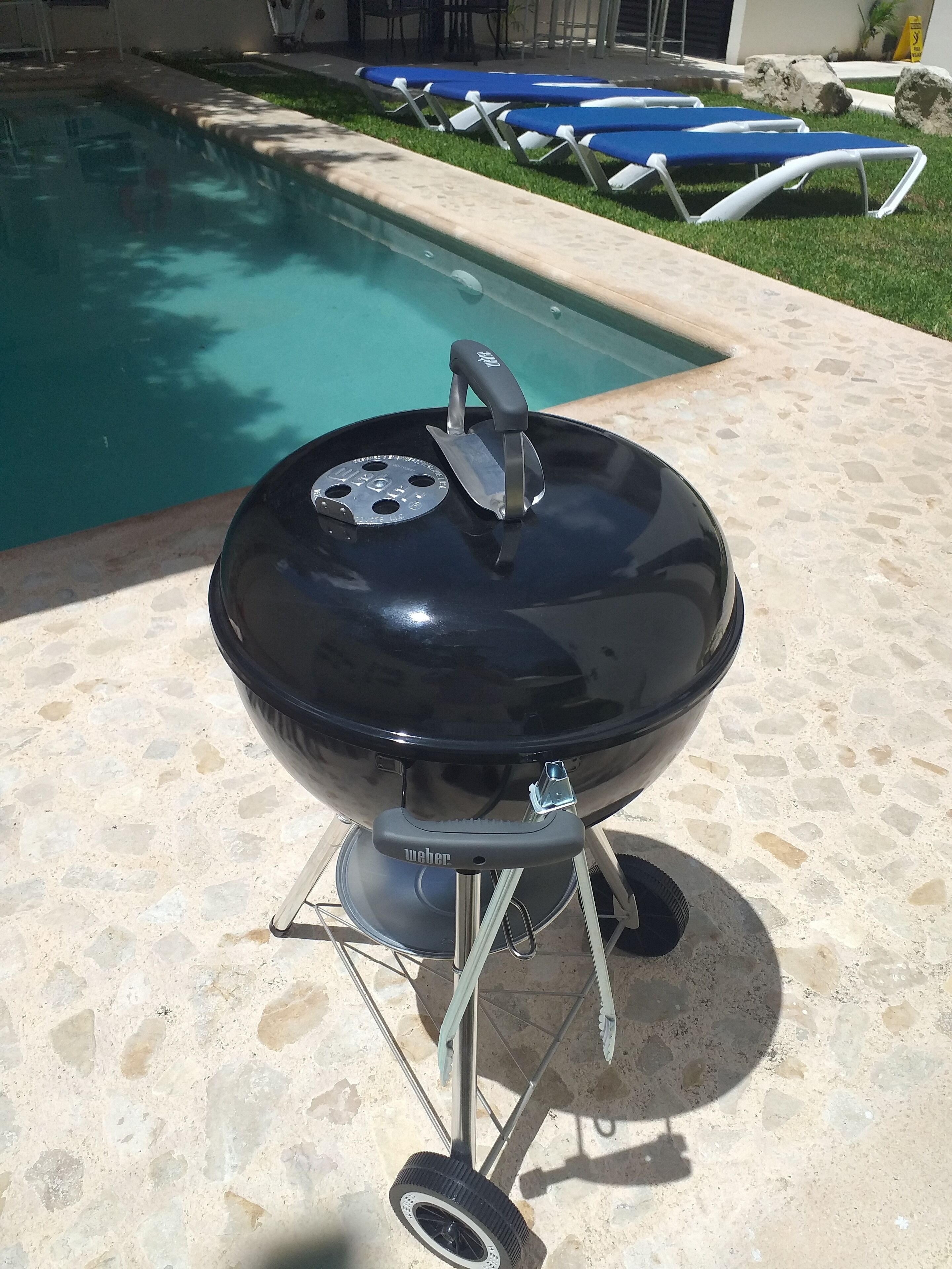 Una parrilla Weber portátil con tapa y asa, ubicada en un patio de piedra cerca de una piscina con sillones de descanso.