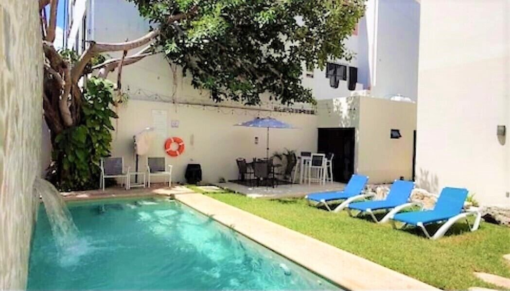 Una piscina con sillas de descanso y una mesa, rodeada de césped y un edificio con puerta y ventanas.