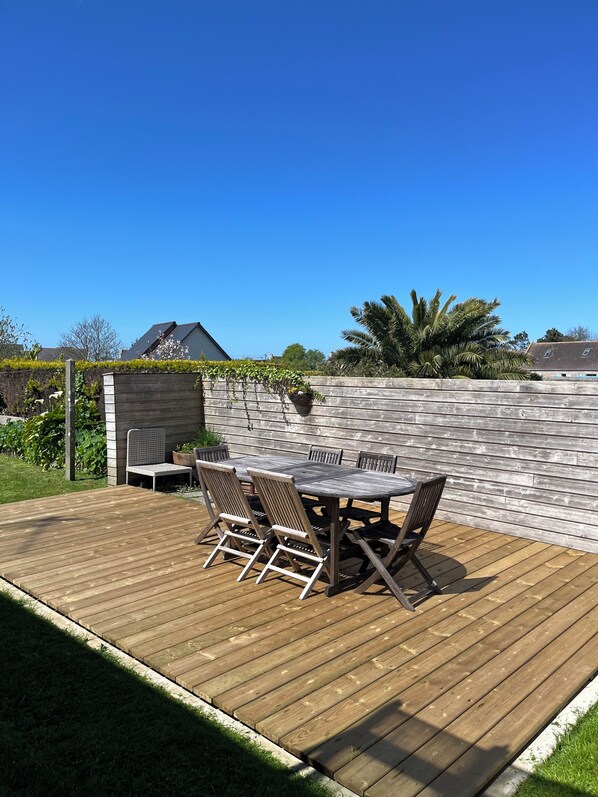 Una terraza de madera con mesa y sillas, rodeada por un muro de piedra y follaje.