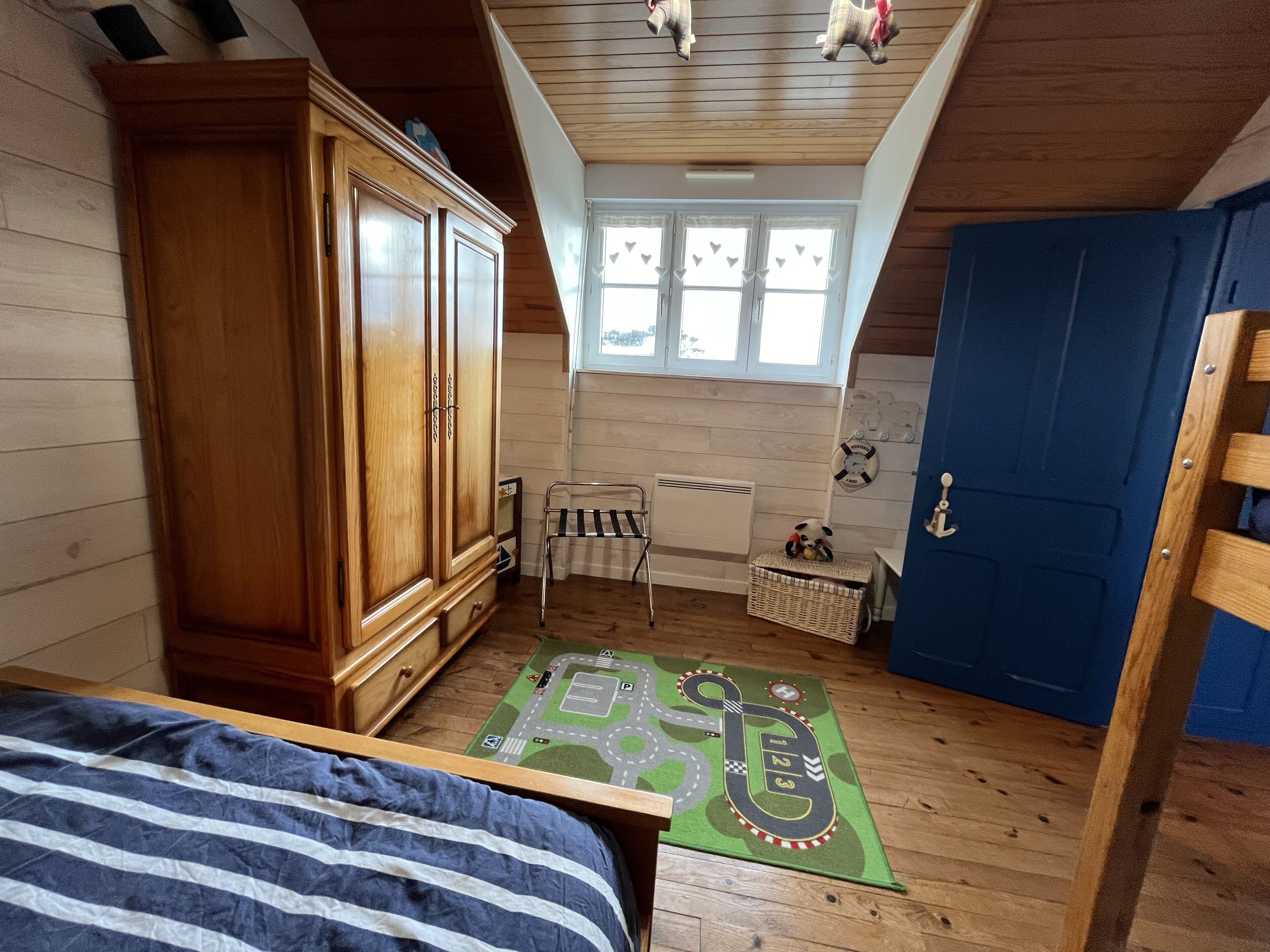 A bedroom with a wooden wardrobe, a bed with a striped blanket, a small table, a radiator, a window with heart decorations, and a blue door.