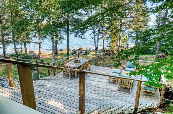 A wooden deck with outdoor furniture, overlooking a body of water and surrounded by trees.