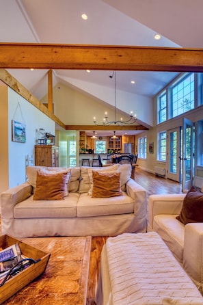 A spacious living room with a high ceiling, a wooden beam structure, a dining area, and large windows.