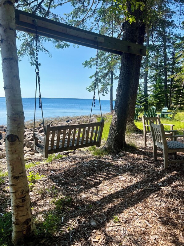 A wooden swing set, a bench, and green Adirondack chairs by a lakeside.