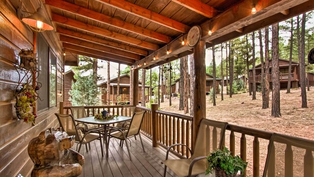 A wooden deck with a table and chairs, surrounded by a wooden railing, overlooking a forested area with cabins. - 3 Mi to Fool Hollow Lake: Romantic Show Low Cabin (Show Low)