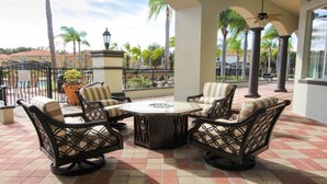 A patio with four cushioned chairs and a central table, surrounded by a tiled floor and palm trees in the background.