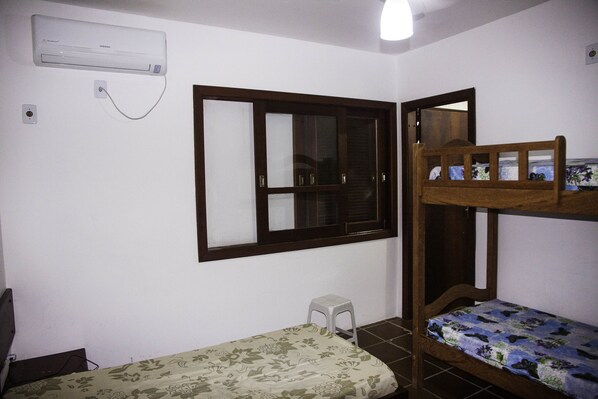 A small room with a bunk bed, a window, a wall-mounted air conditioner, and a small stool.