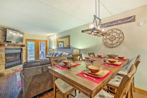 Uma sala de jantar com uma mesa de madeira posta para seis pessoas, rodeada por cadeiras de madeira. Uma lareira de pedra é visível à esquerda, e um relógio de parede grande está pendurado acima da mesa.