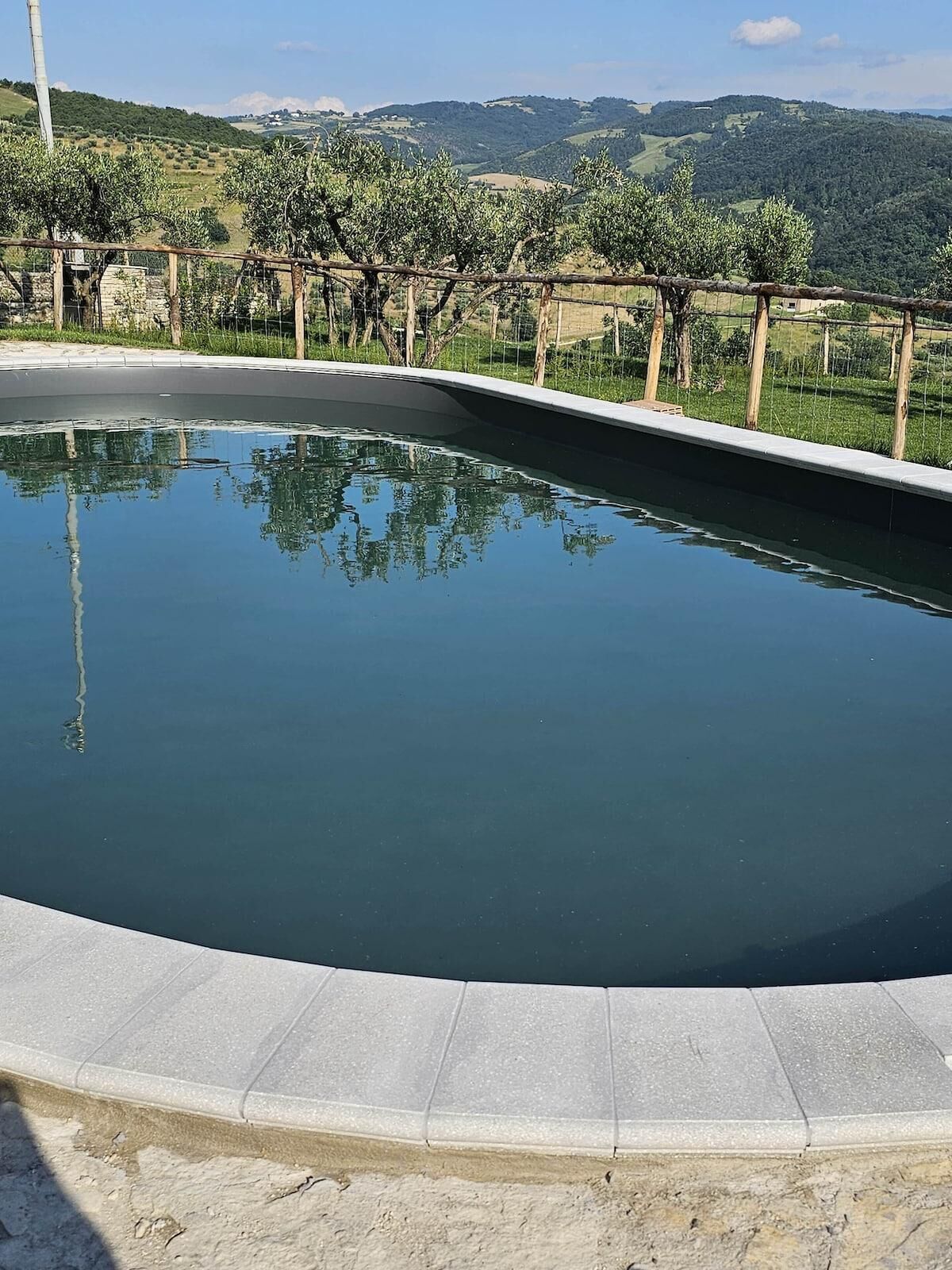 Una piscina con borde de piedra, rodeada de valla de madera y árboles, con vistas a colinas al fondo.