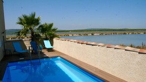 A swimming pool with a wooden deck, lounge chairs, and a view of a body of water and distant hills. - House / Villa - Frontignan (Frontignan)