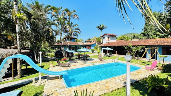 Une piscine avec un toboggan, entourée de palmiers et surplombée d’un ciel bleu azur.