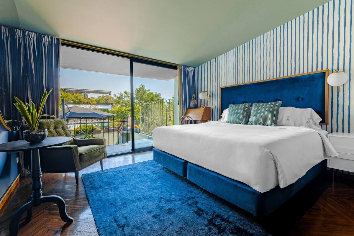 Une chambre avec un grand lit, un bureau et un balcon offrant une vue sur la verdure et un immeuble.