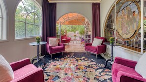 Een kamer met een groot bloementapijt, twee roze fauteuils, een ronde tafel en uitzicht op een tuin door een glazen deur.