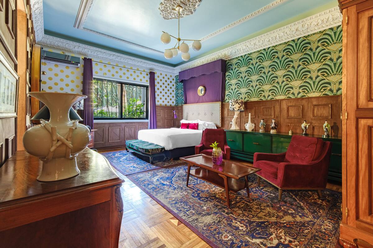 Une pièce comprenant un lit, un fauteuil rouge, une armoire en bois et un grand vase posé sur une table.