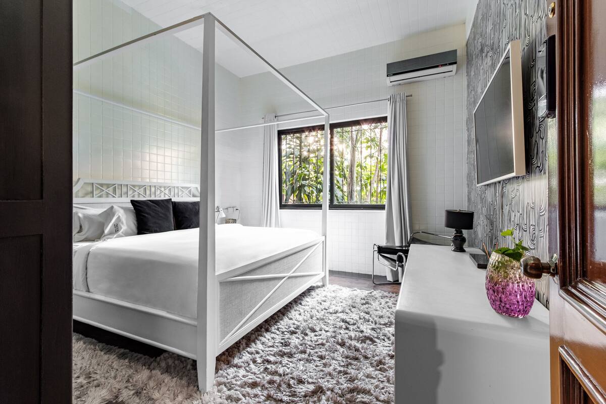 Une chambre moderne avec un lit à baldaquin, une grande fenêtre avec des rideaux, un téléviseur à écran plat fixé au mur et un petit bureau agrémenté d’un vase de fleurs.