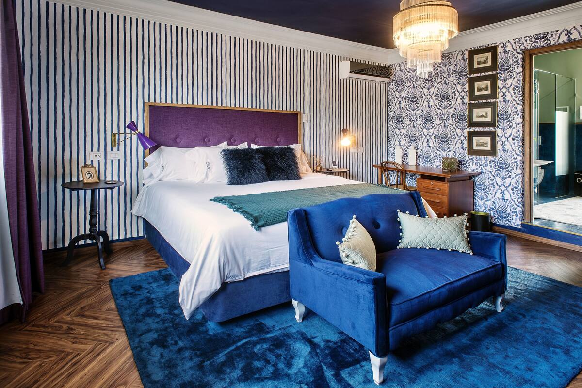 Une chambre à coucher avec un grand lit, un fauteuil bleu, une table de chevet en bois et un lustre.