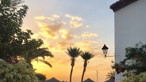 A pathway leading to a building with a stone gate, palm trees, and a sunset in the background.