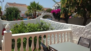 A white balcony with a table and chair, surrounded by lush greenery and a pool.