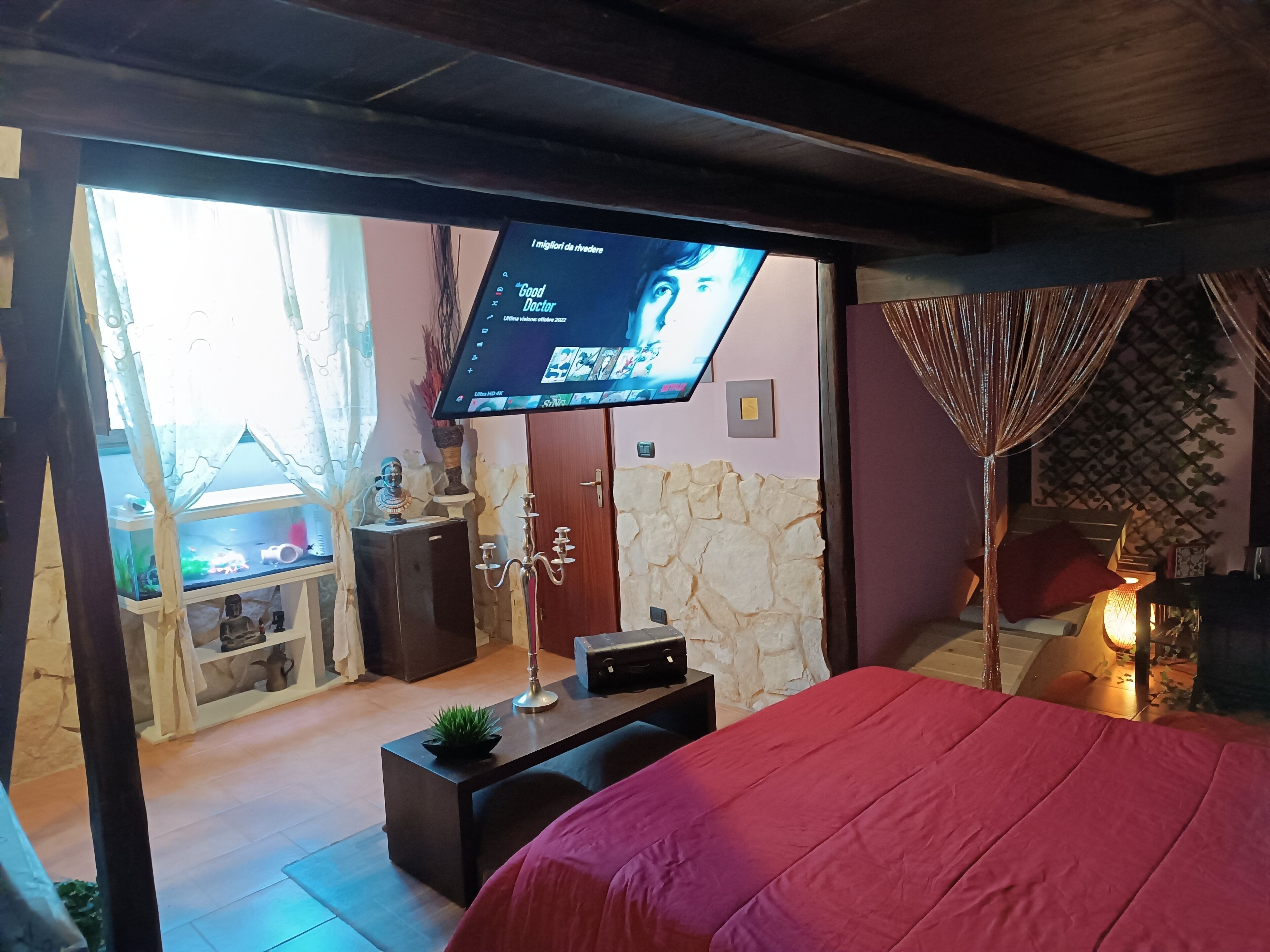 A bedroom with a bed, a TV mounted on a wall, a stone wall, and a small table with a plant.