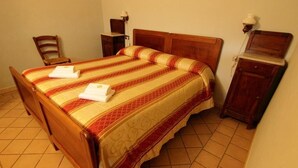 Uma cama bem arrumada com colcha listrada e duas toalhas, uma mesinha de cabeceira de madeira com abajur, e uma cadeira em um quarto com piso de azulejos.