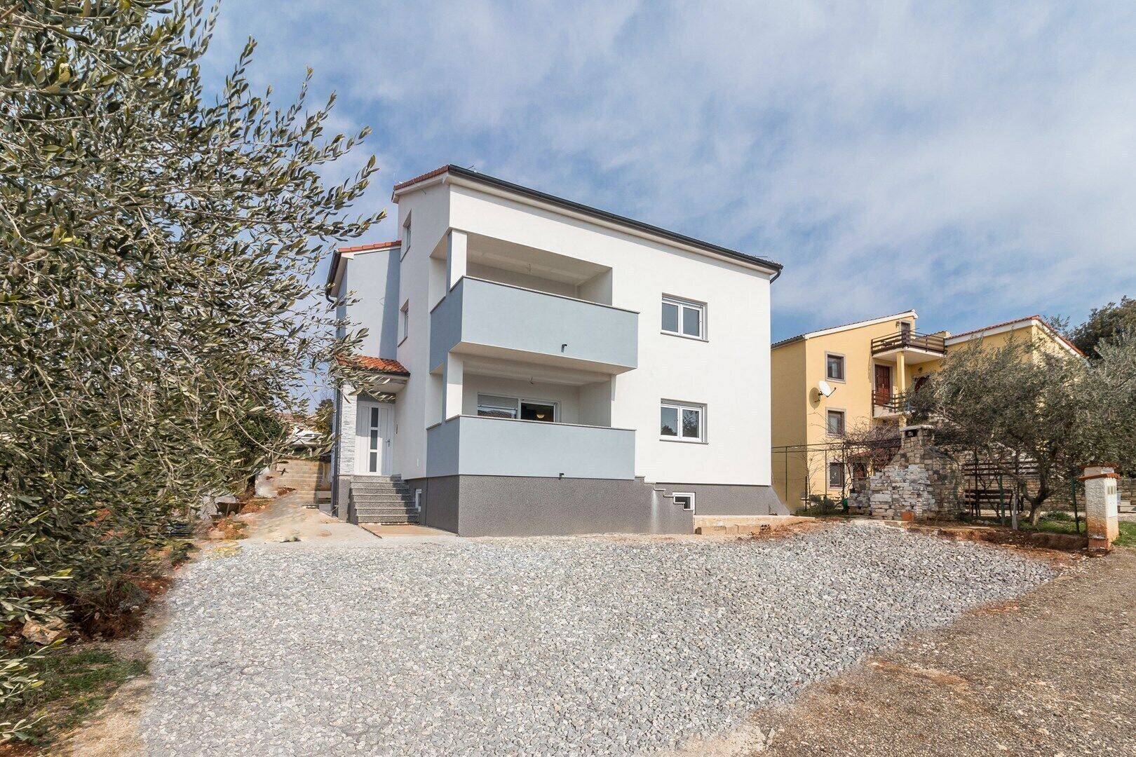 A modern two-story residential building with a flat roof, a balcony on the second floor, and a gravel driveway leading to the entrance.