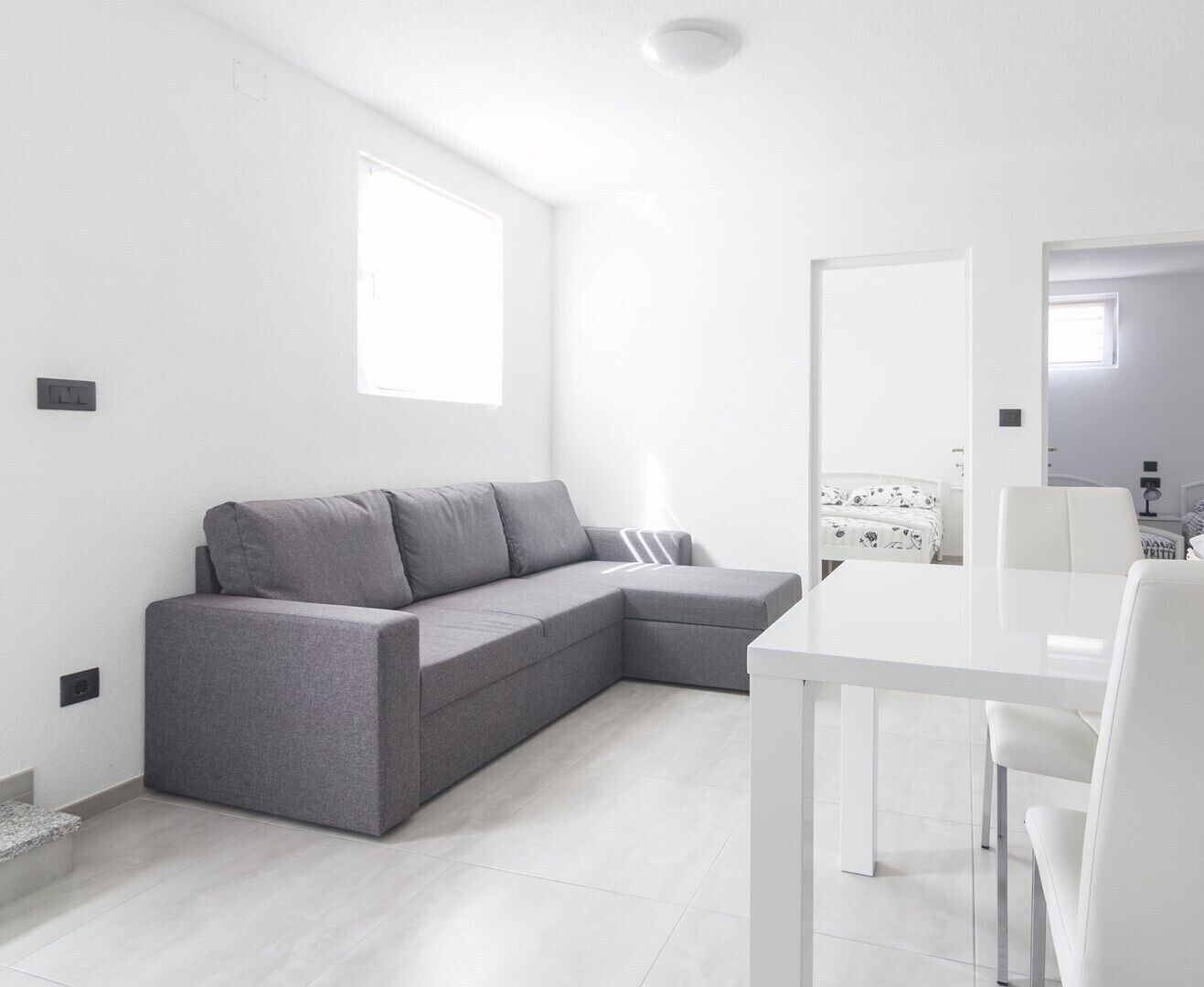 Una sala de estar moderna y minimalista con un sofá seccional gris, una mesa de comedor blanca y una pequeña área de cocina.