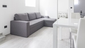 Ein modernes, minimalistisches Wohnzimmer mit einer grauen Eckcouch, einem weißen Esstisch und einer kleinen Küchenzeile.