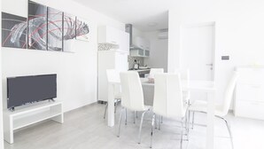 Ein modernes, weißes Wohnzimmer mit einem Flachbildfernseher, einem Essbereich mit weißen Stühlen und einem Tisch sowie einer Küche im Hintergrund.