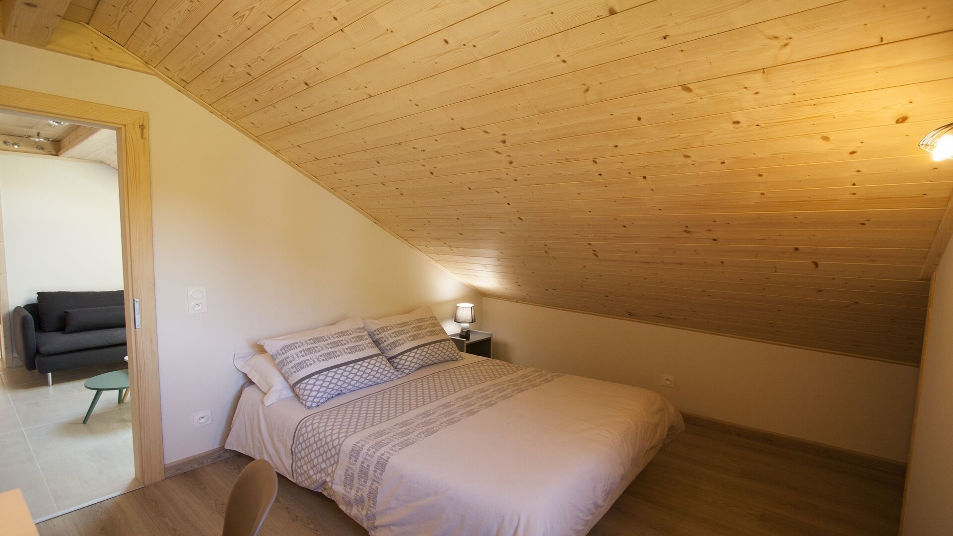 Un dormitorio con techo inclinado, una cama con ropa de cama estampada, una lámpara de noche y un sofá en la habitación contigua.