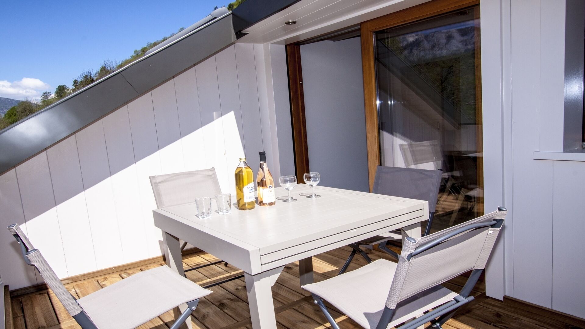 Een witte tafel met twee stoelen op een houten terras, een fles wijn en twee wijnglazen.