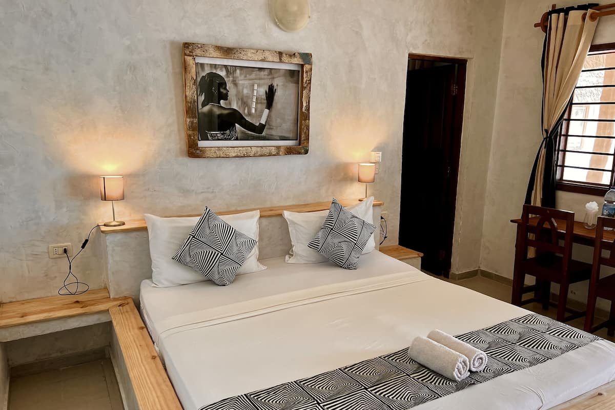 Una cama bien hecha con almohadones estampados, una mesita de noche con lámpara y un cuadro en la pared.