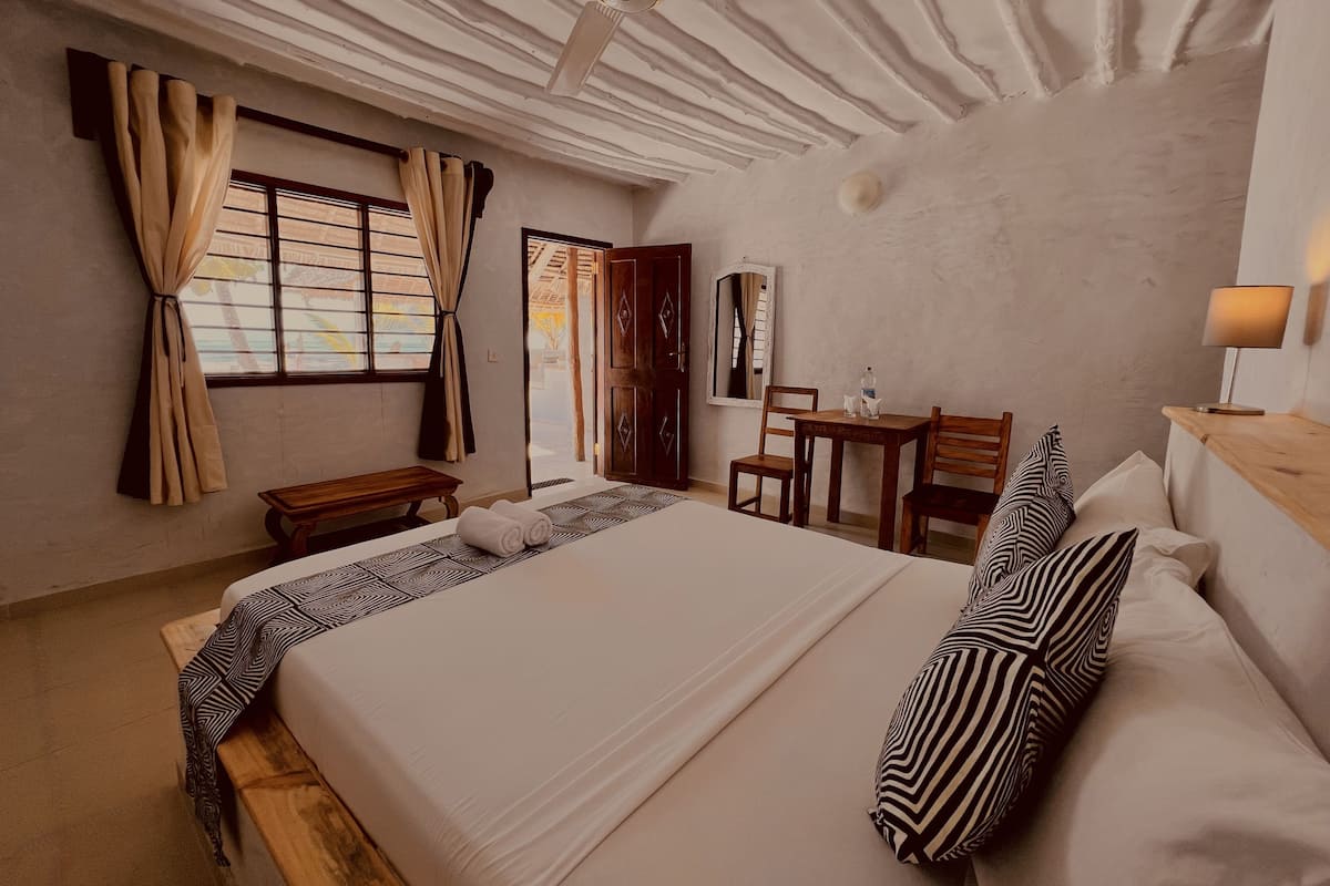Habitación de hotel con una cama grande, un escritorio y una silla. Hay un ventilador de techo, una ventana con cortinas y un espejo en la pared.
